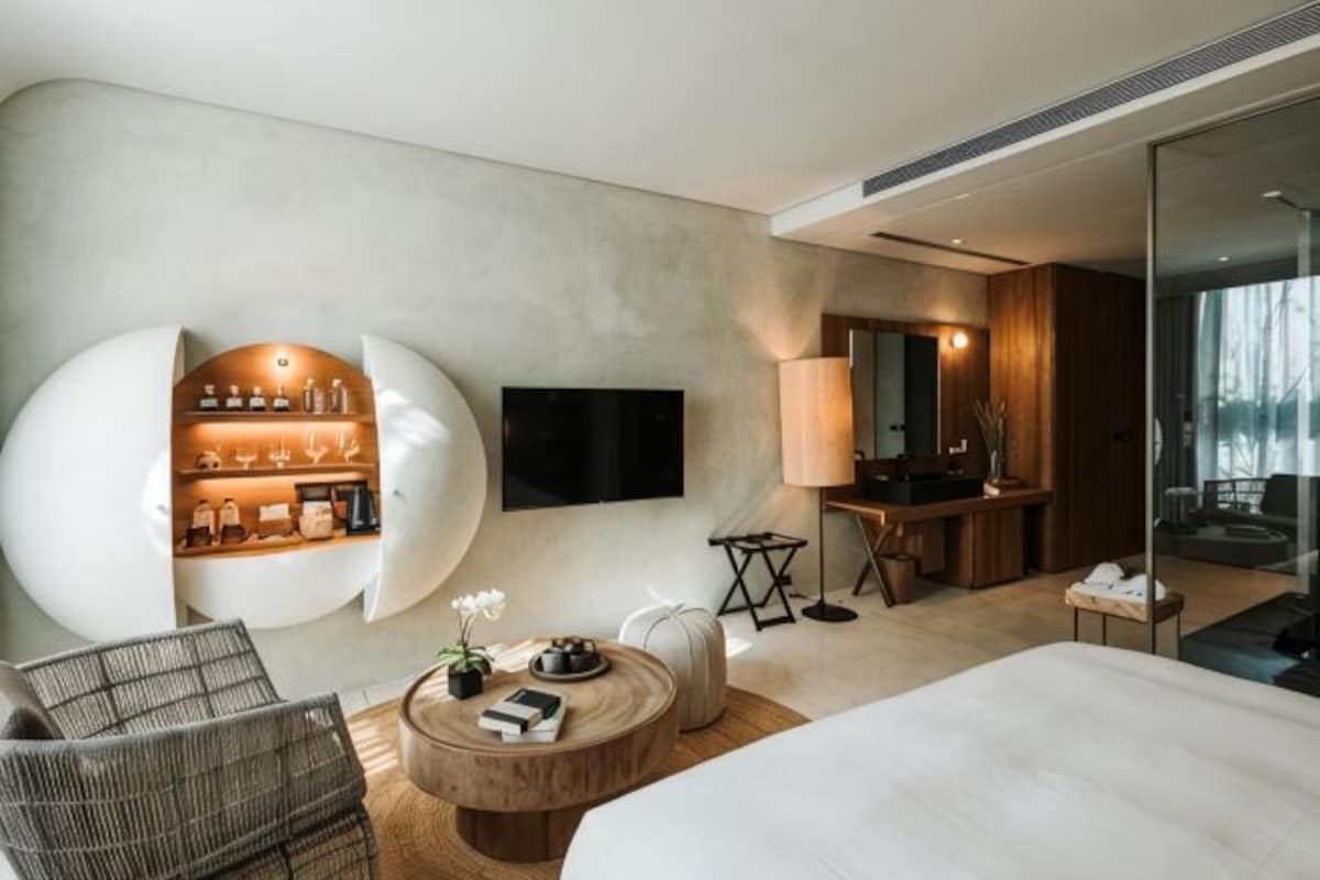 A modern hotel room with a large bed, a round coffee table, a seating area with a chair, a TV mounted on the wall, and a bathroom with a mirror and sink.