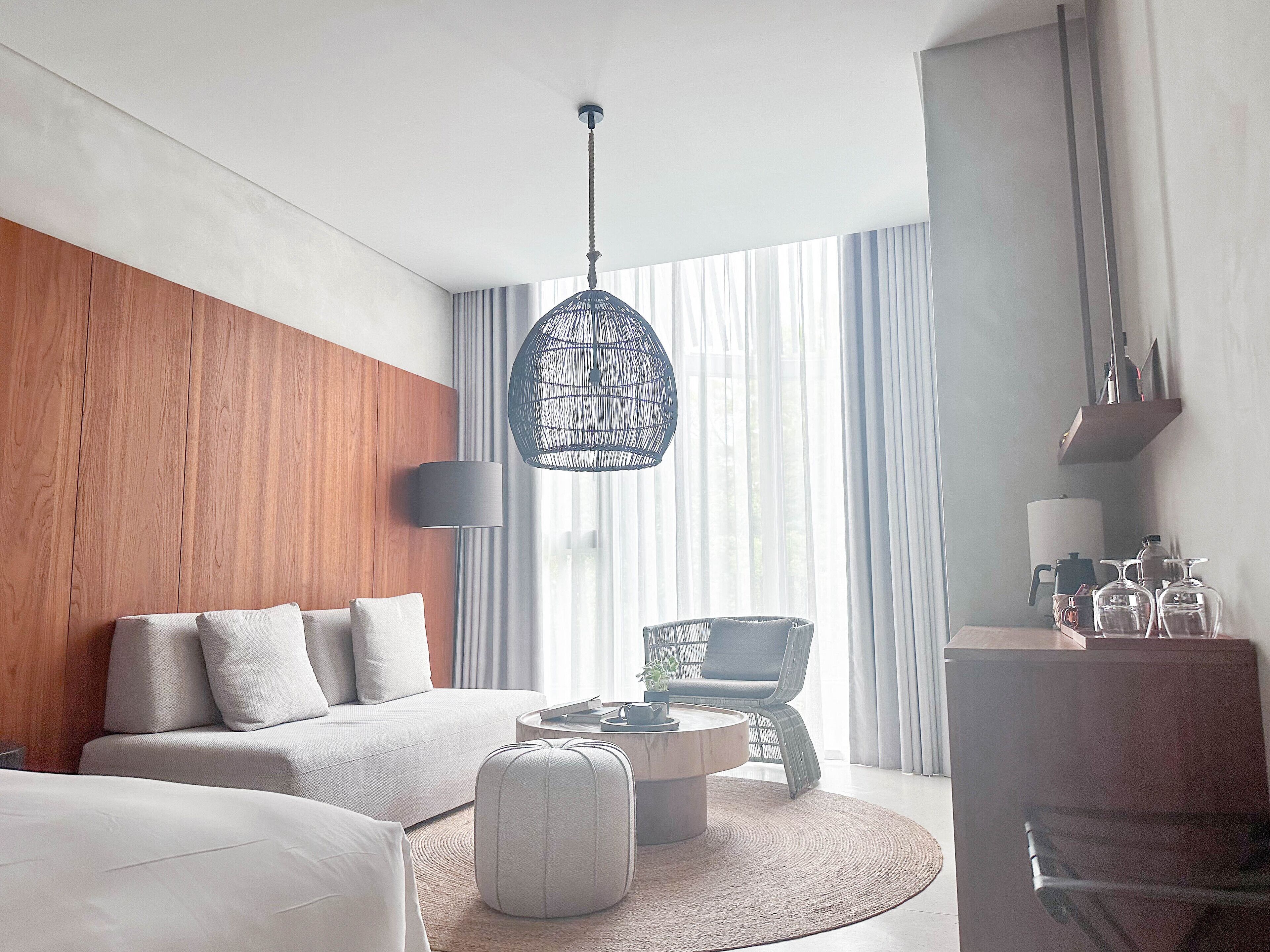 Quarto de hotel moderno com parede de destaque em madeira, mesa de centro redonda, sofá, poltrona e luminária pendente.