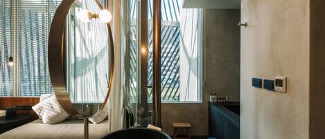 A modern room with a large circular mirror, a stone basin, a bed with a view of a building, and a small wooden stool.
