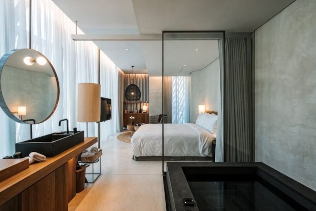 A modern hotel room with a large bed, a separate bathroom with a bathtub, a vanity with a mirror, and a view through a sliding door.