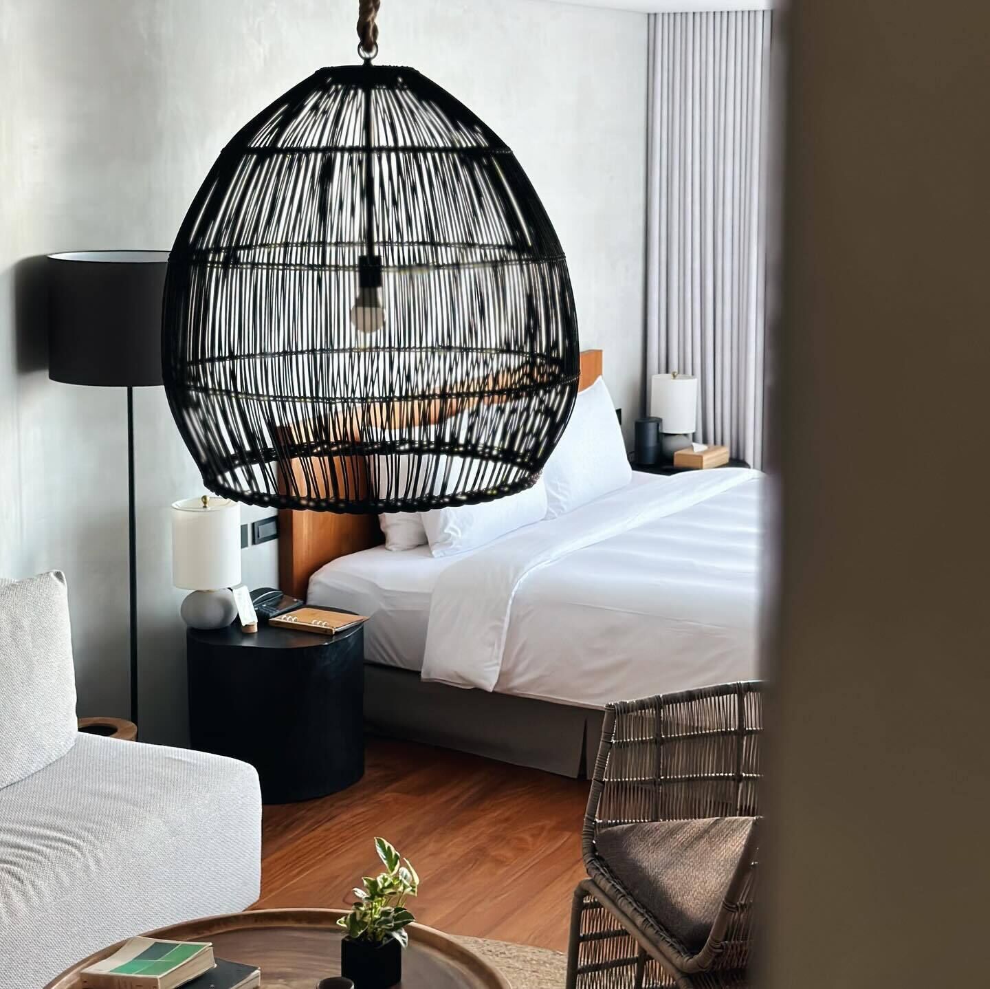 Ein modernes Hotelzimmer mit einem großen Bett, einem hängenden Rattan-Sessel, einem runden Beistelltisch mit Lampe und einer kleinen Pflanze.
