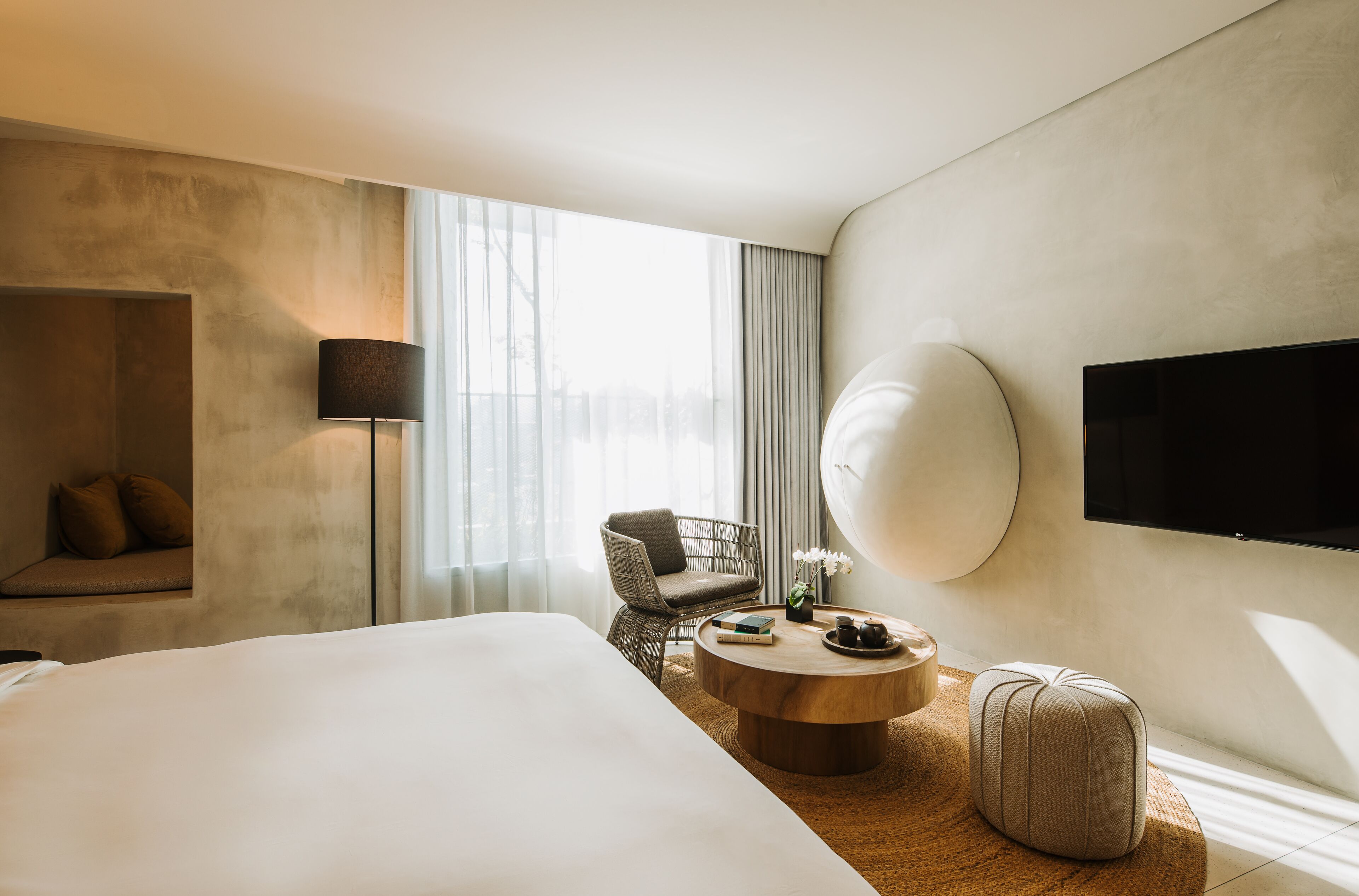 Quarto de hotel moderno com uma cama grande, uma mesa de centro redonda de madeira, uma poltrona confortável, uma pequena mesa lateral com abajur e uma TV de tela plana na parede.