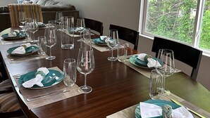 A dining area with a long wooden table set for a meal, surrounded by chairs, with a clock on the wall and a window with a view of greenery. - Great Home in South County, Close to Narraganstt and Newport &.08 miles from URI (South Kingstown)
