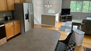 A modern kitchen with wooden cabinets, a stainless steel refrigerator, and a built-in island. There is a dining area with a table and a sofa. - Great Home in South County, Close to Narraganstt and Newport &.08 miles from URI (South Kingstown)