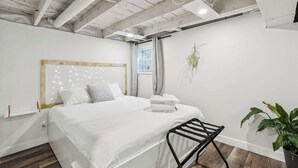 A modern bedroom with a white bed, a wooden headboard, a black metal chair, and a potted plant. - Trendy Kensington | Espresso Maker| Garage| AC (Calgary)