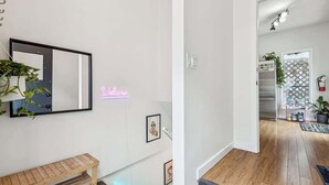 A modern interior with a wooden bench, a neon 'Welcome' sign, and a hallway leading to a kitchen area with a refrigerator and potted plants. - Trendy Kensington | Espresso Maker| Garage| AC (Calgary)
