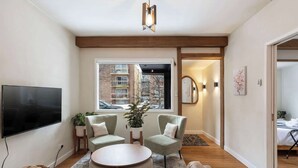 A modern living room with a flat-screen TV, two armchairs, a round coffee table, and a view of a cityscape through a large window. - Trendy Kensington | Espresso Maker| Garage| AC (Calgary)