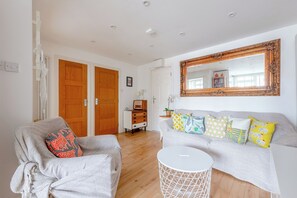 Una sala de estar con un sofá, un sillón y una mesita redonda blanca. Se ven puertas de madera y un armario blanco al fondo.