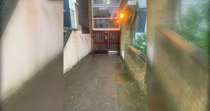 A narrow alleyway with a brick wall on one side and a white wall on the other, leading to a gated entrance.