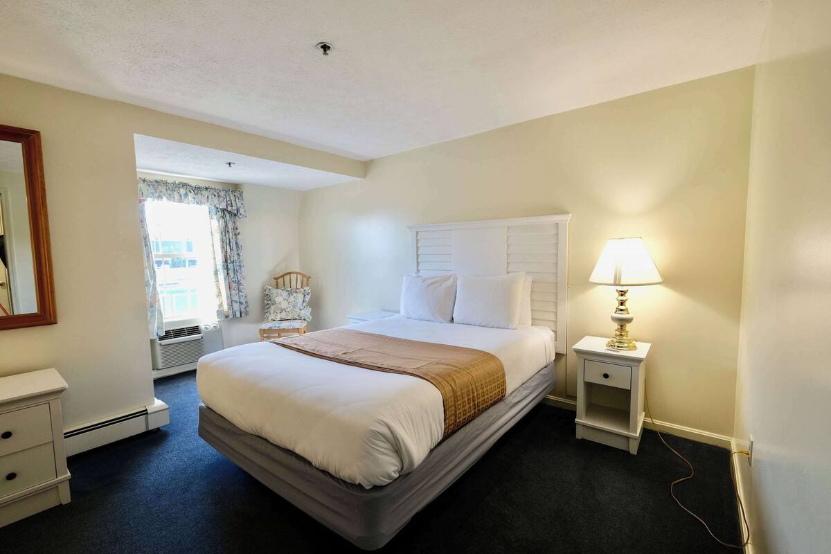 A hotel room with a large bed, a bedside lamp, a nightstand, a dresser, and a window with curtains.