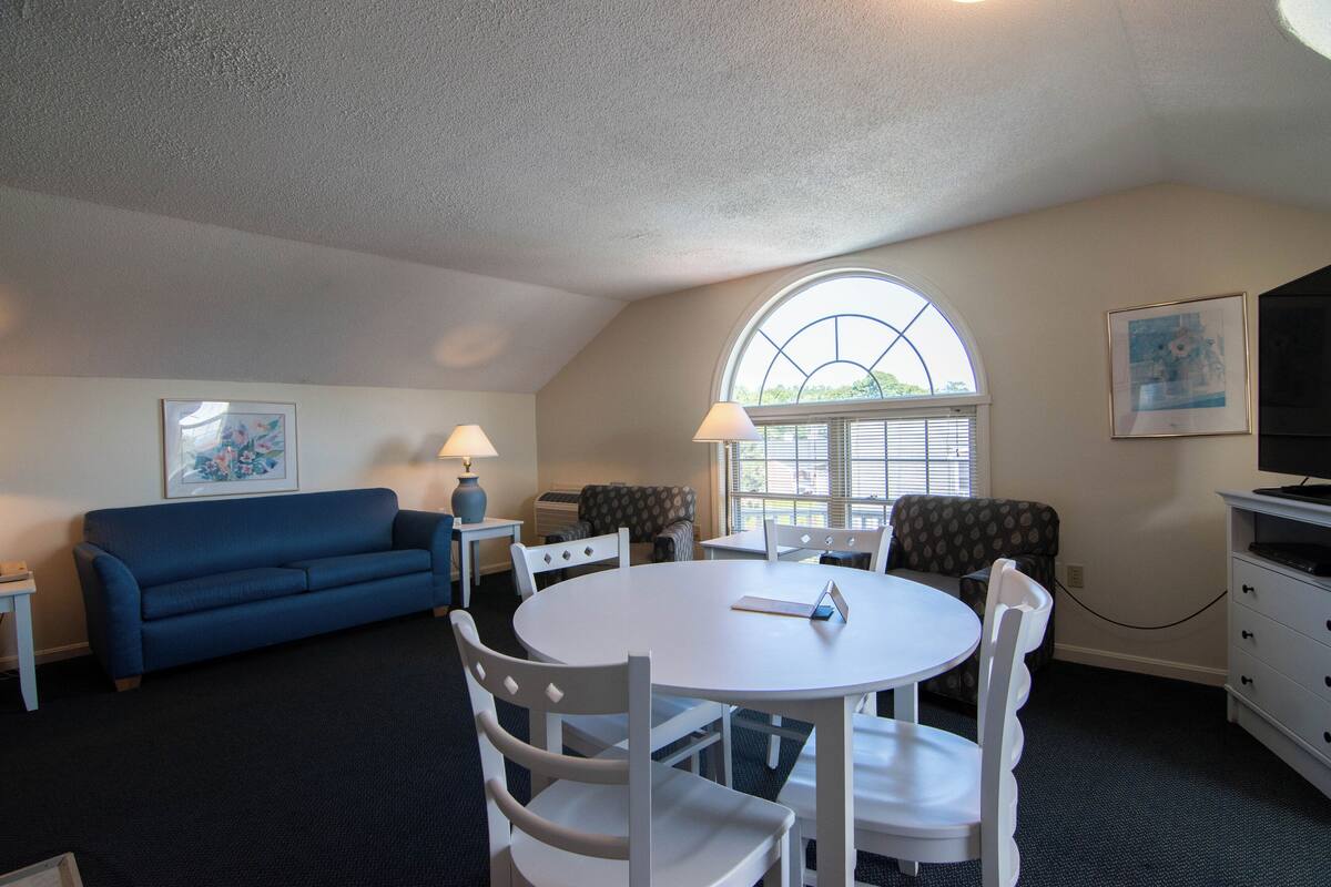 A room with a round white table, white chairs, a blue sofa, a TV, and a window with blinds.