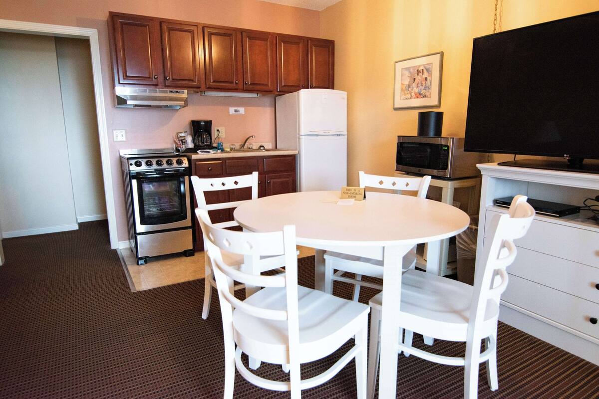 A compact kitchen with wooden cabinets, a white refrigerator, a stove, and a microwave. There is a dining table with four chairs and a sign indicating a kitchen area.