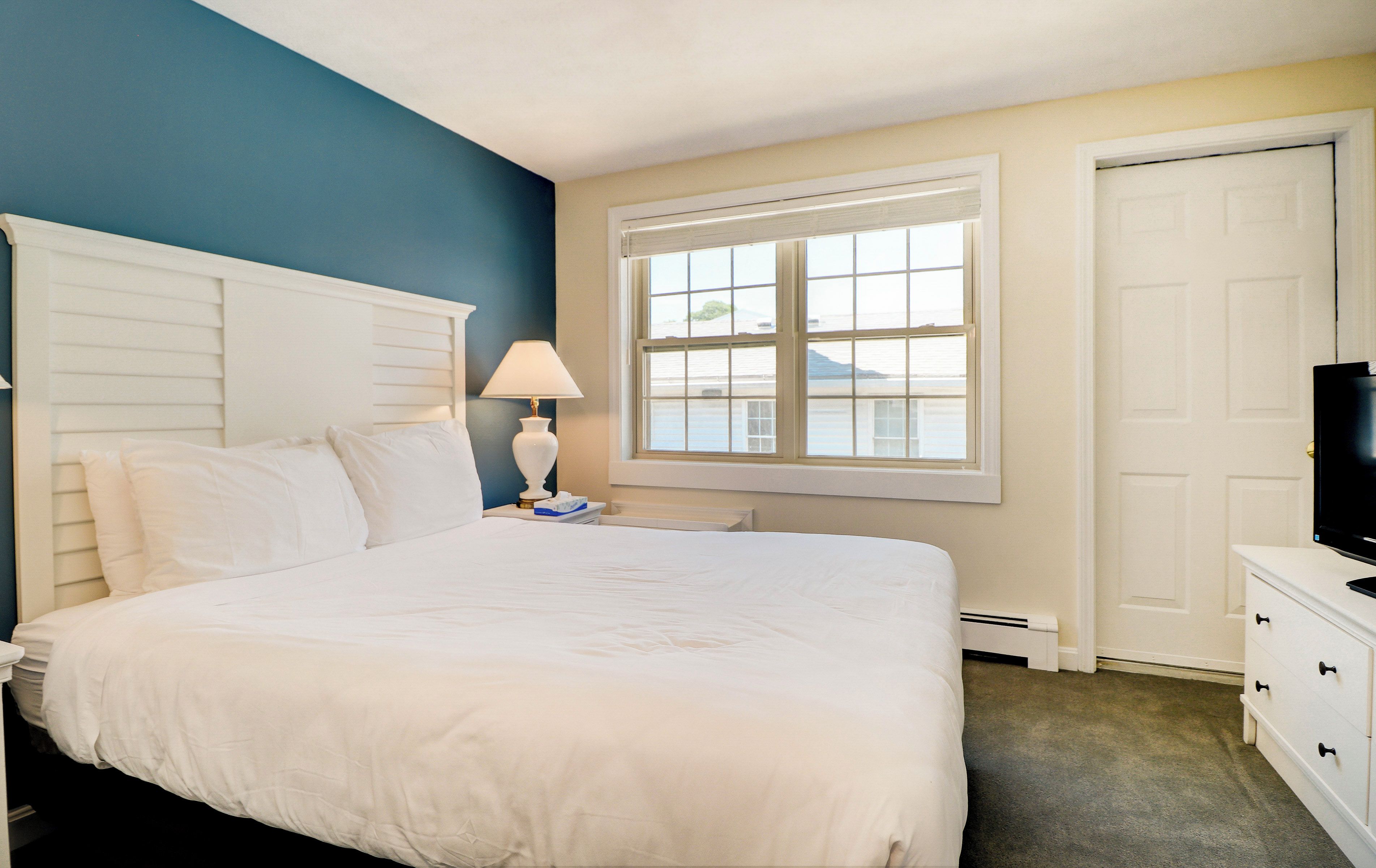 A hotel room with a large bed, a bedside lamp, a window with blinds, a door, and a dresser with a television.