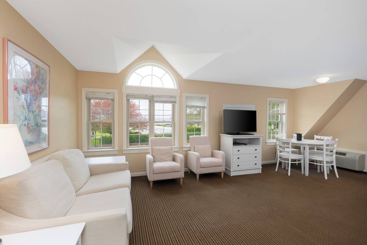 A spacious living room with a high ceiling, a large window, a flat-screen TV, a dining table with chairs, a sofa, and a cabinet.