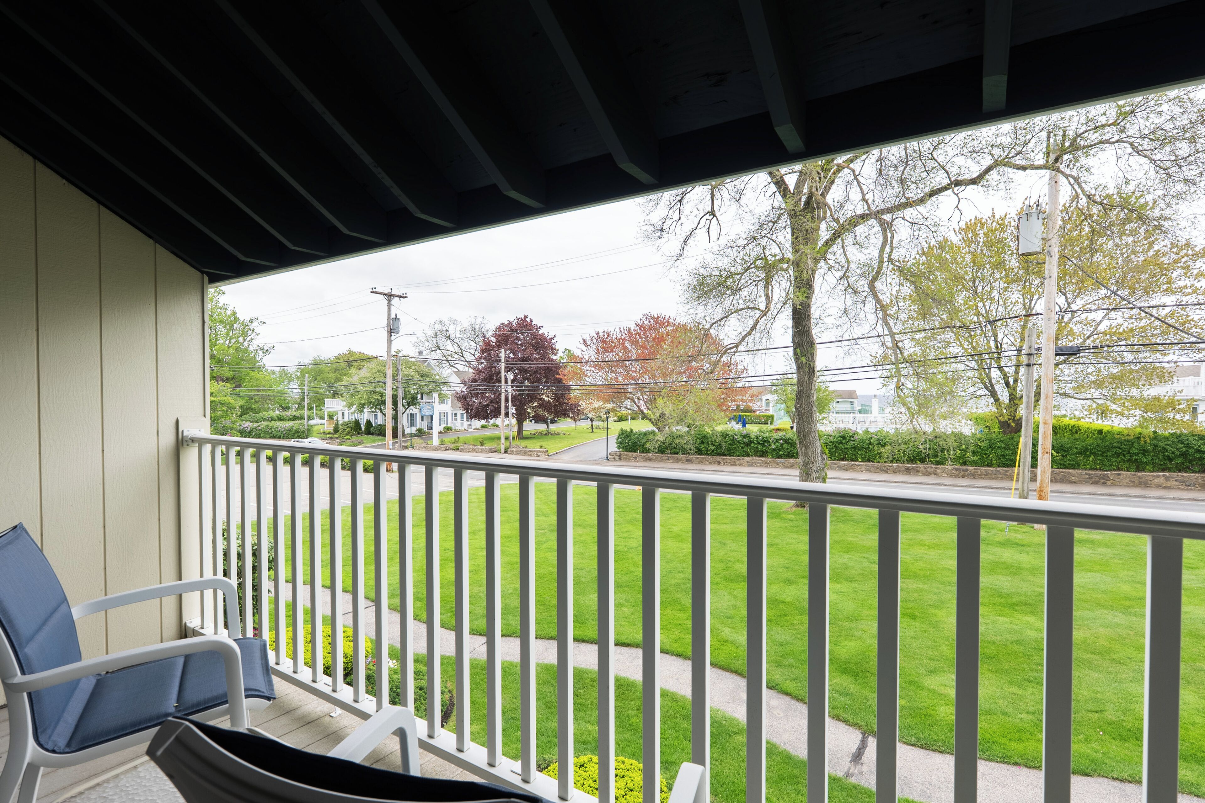 A balcony with a white railing, a blue chair, and a view of a green lawn and a street with trees and buildings.