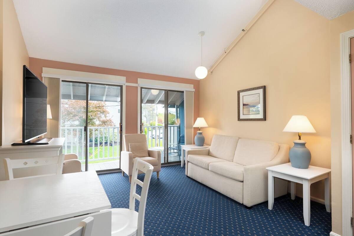 A hotel room with a white desk, a white sofa, a small table with a lamp, and a chair. There is a large window with a view of the outdoors.