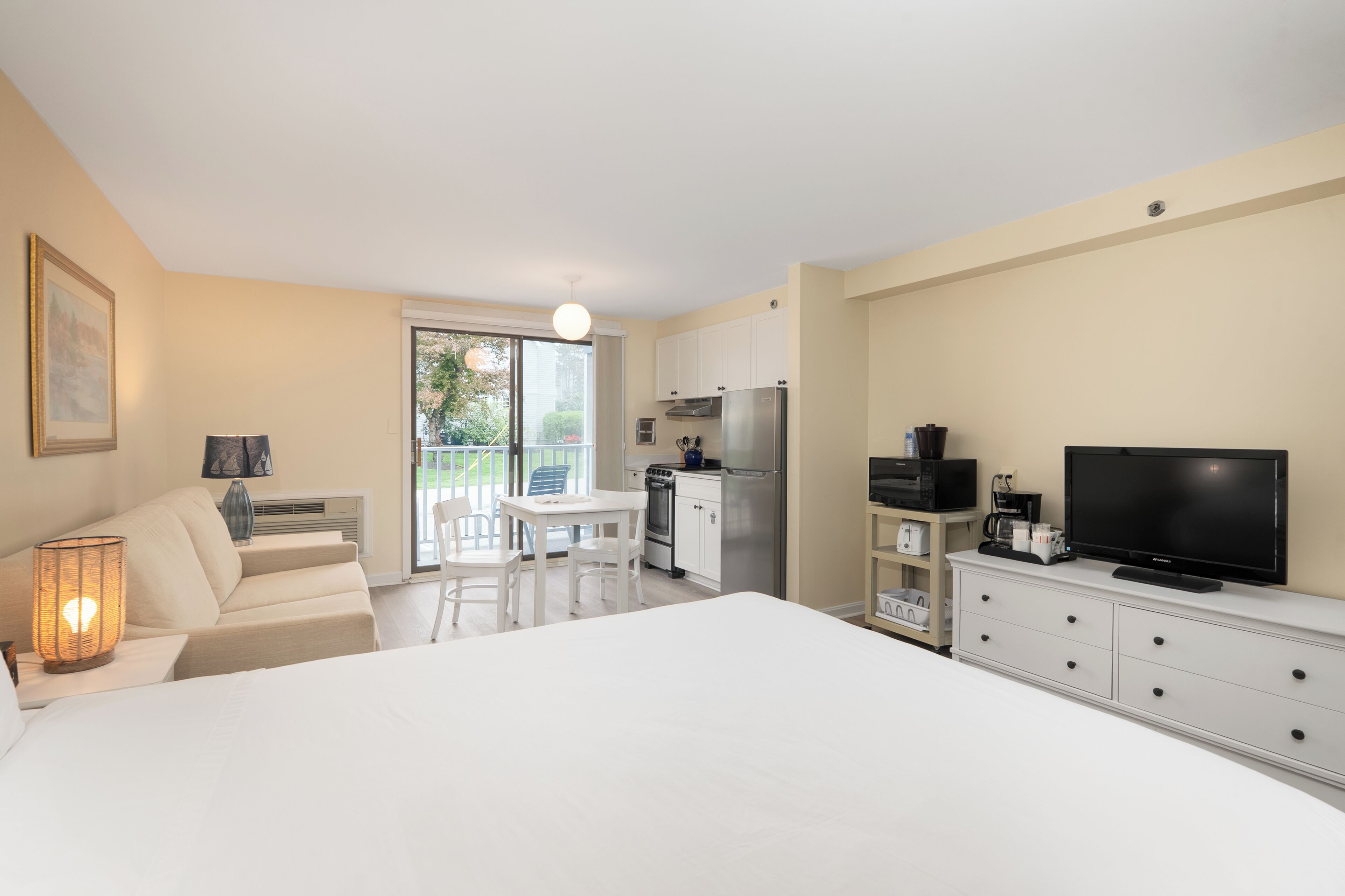 A modern hotel room with a large bed, a dining area, kitchenette, and a TV.