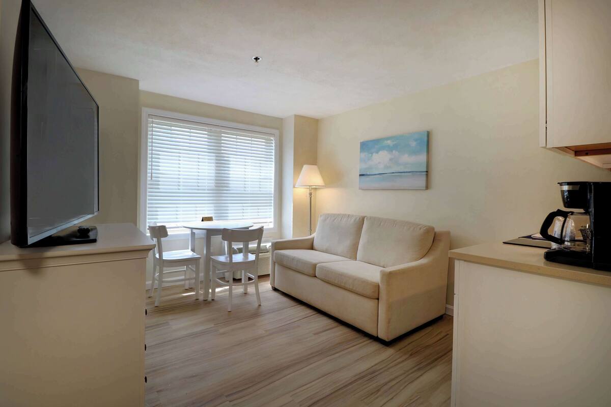 A compact living space with a sofa, a small dining table with chairs, a television, a lamp, and a coffee maker.
