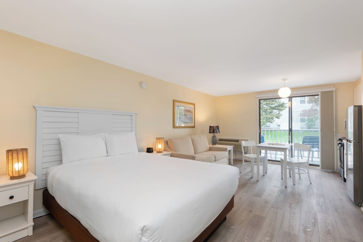 A hotel room with a large bed, a desk, a chair, a lamp, and a refrigerator.