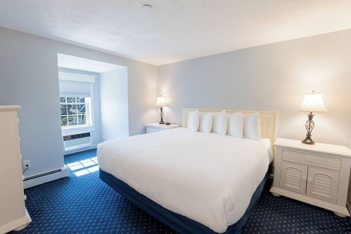 A hotel room with a large bed, two bedside tables with lamps, a window with blinds, and a blue carpet.