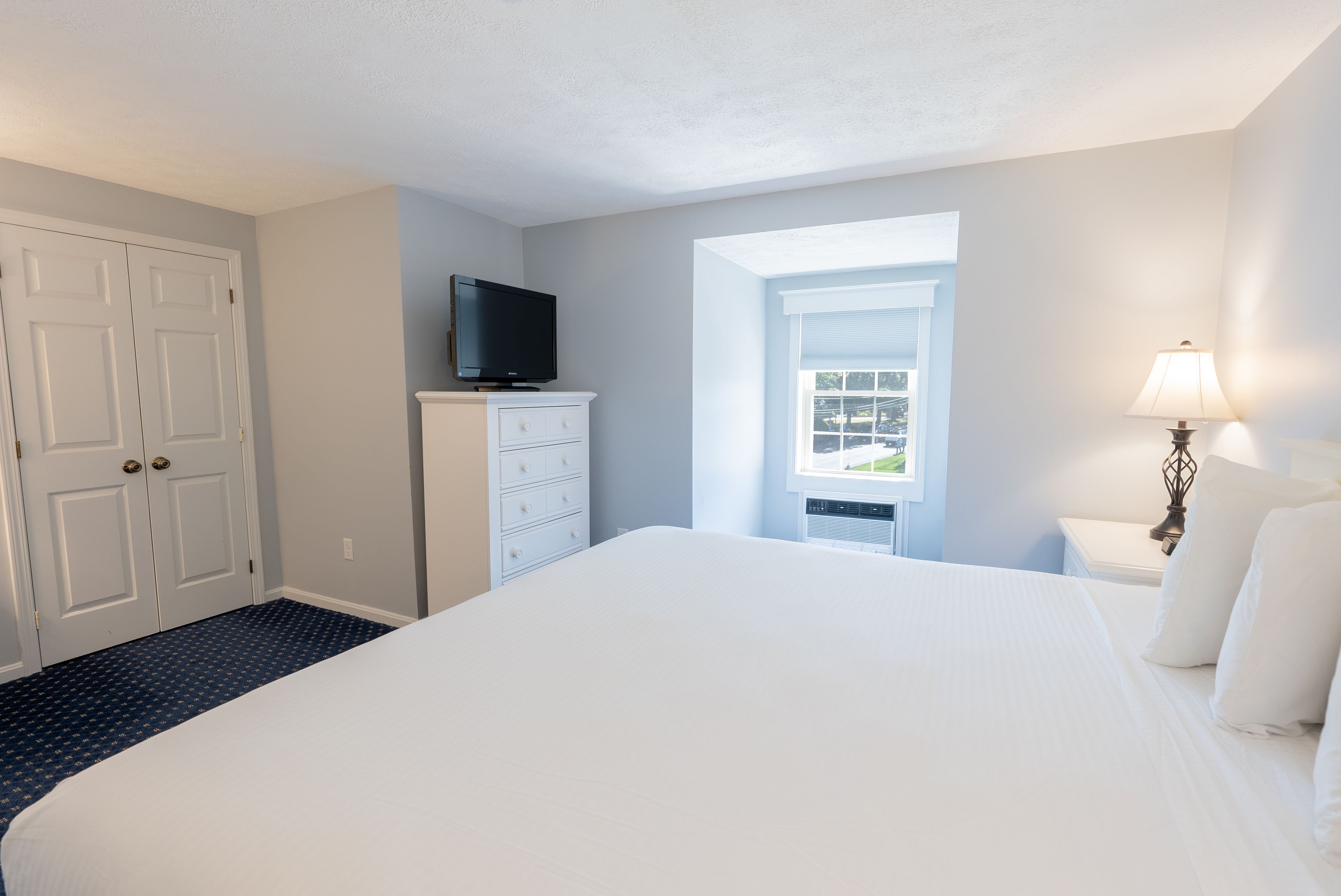 A hotel room with a large bed, a television on a dresser, a window with blinds, and a nightstand with a lamp.