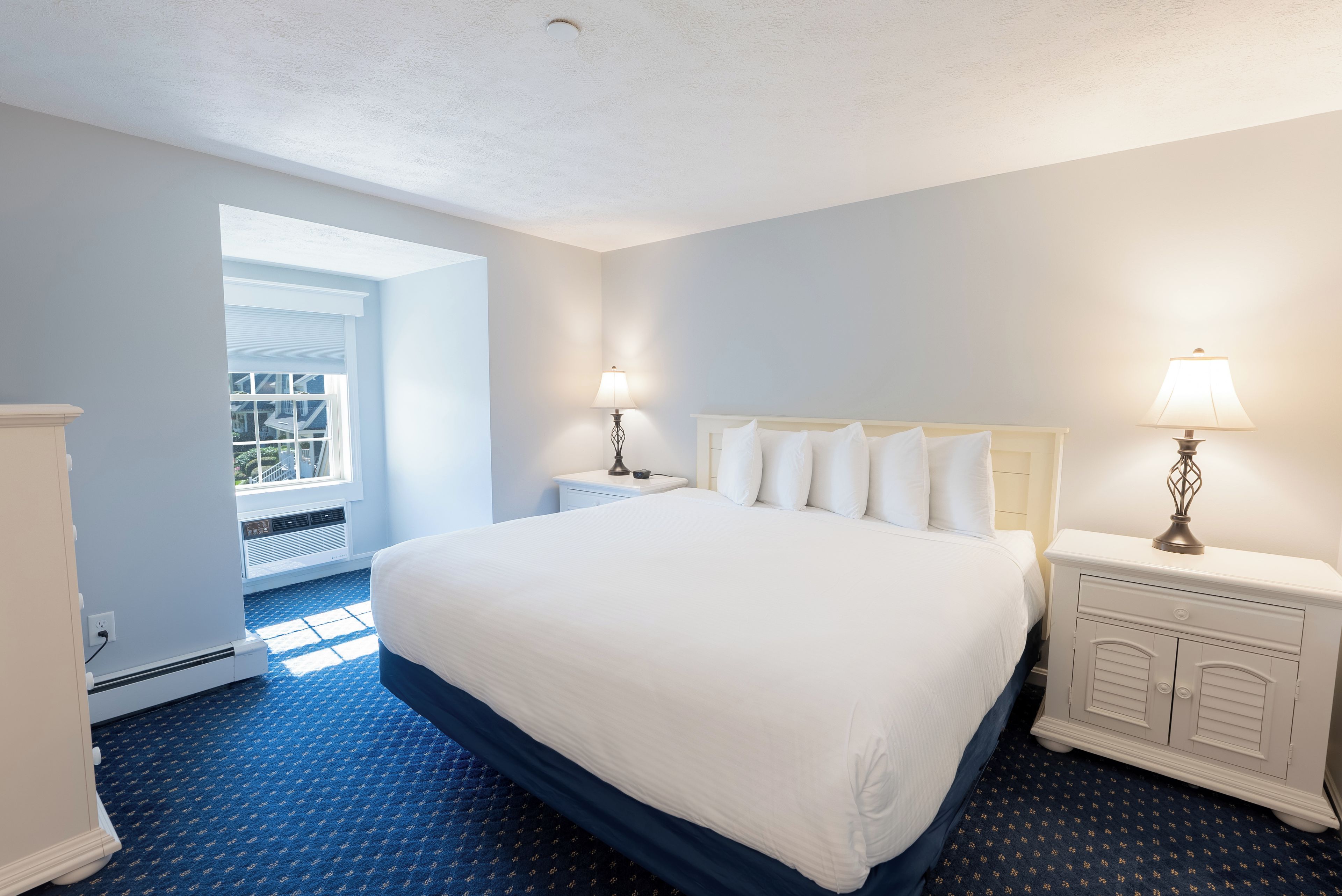 Une chambre d’hôtel avec un grand lit, deux tables de chevet avec des lampes, une fenêtre avec des stores et une moquette bleue.