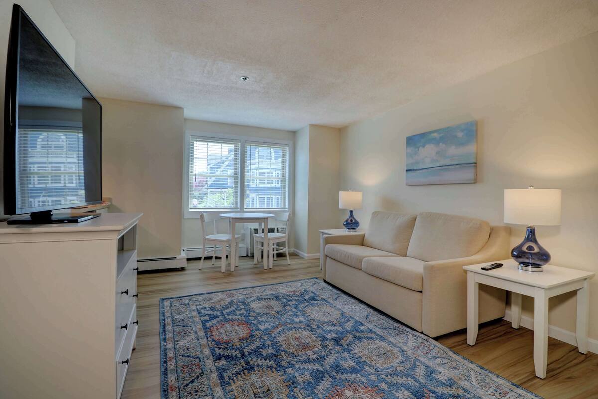 A living room with a flat-screen TV, a beige sofa, two side tables with lamps, a patterned rug, and a dining area with a table and chairs.