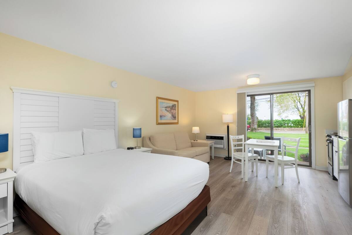 A hotel room with a large bed, a sofa, a dining table with chairs, a refrigerator, and a window with a view of the outdoors.