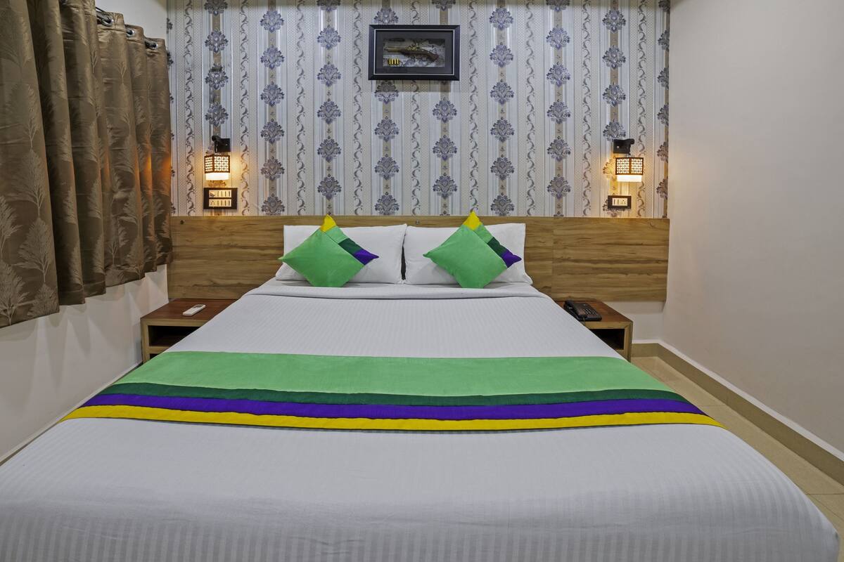 Cama bem arrumada com roupa de cama verde e roxa, duas mesas de cabeceira com luminárias e papel de parede estampado.
