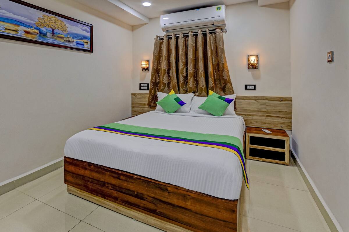 Quarto de hotel com cabeceira de madeira, cama com roupa de cama branca e almofadas coloridas, mesa de cabeceira com controle remoto e ar condicionado acima da cama.