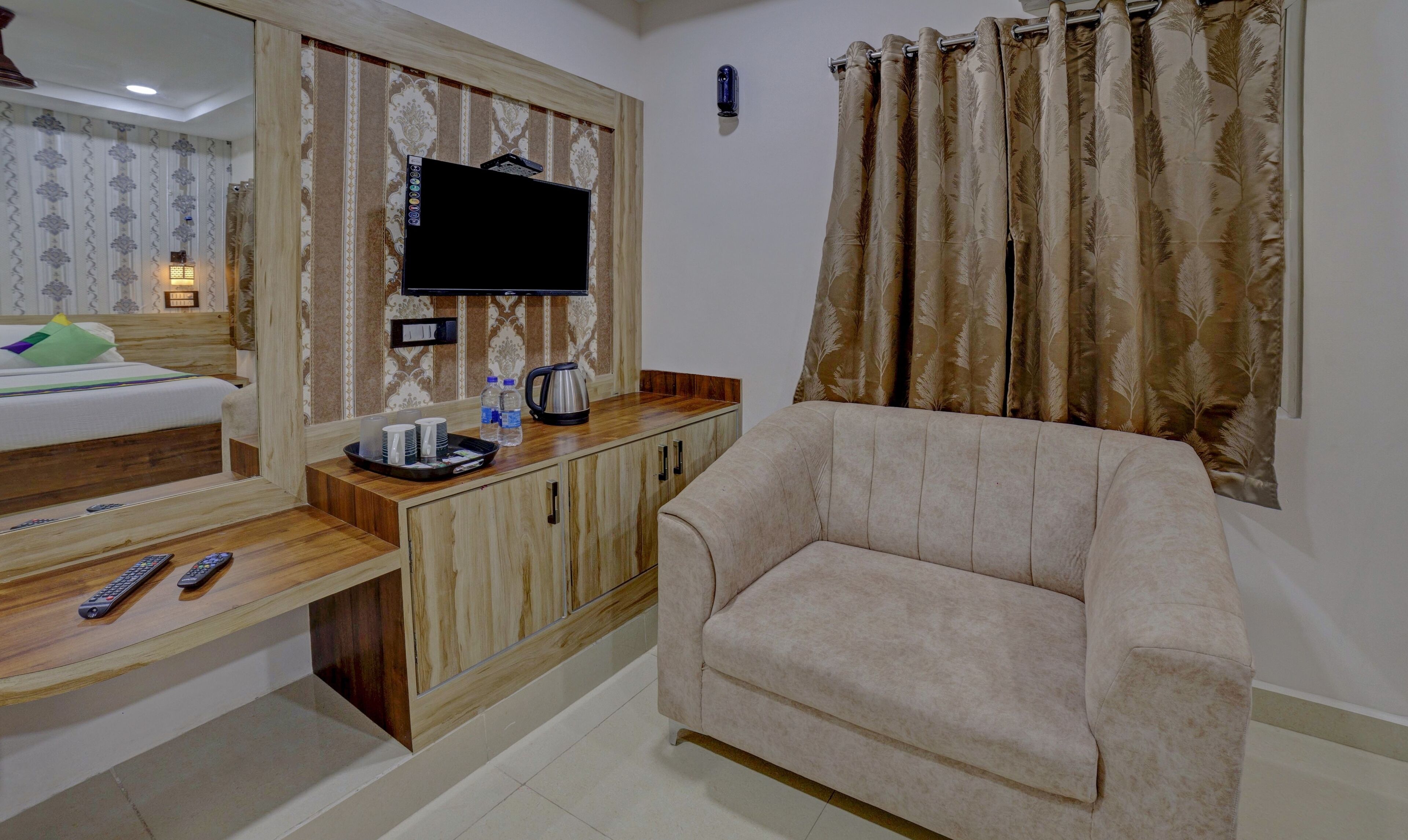A hotel room with a bed, a TV mounted on the wall, a sofa, a wooden cabinet, and a window with curtains.