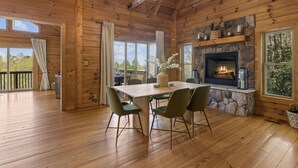 A wooden cabin interior with a stone fireplace, a dining table with chairs, and a large window overlooking a deck. - Pisgah Paradise • Private • Game Room • Large Deck (Pisgah Forest)