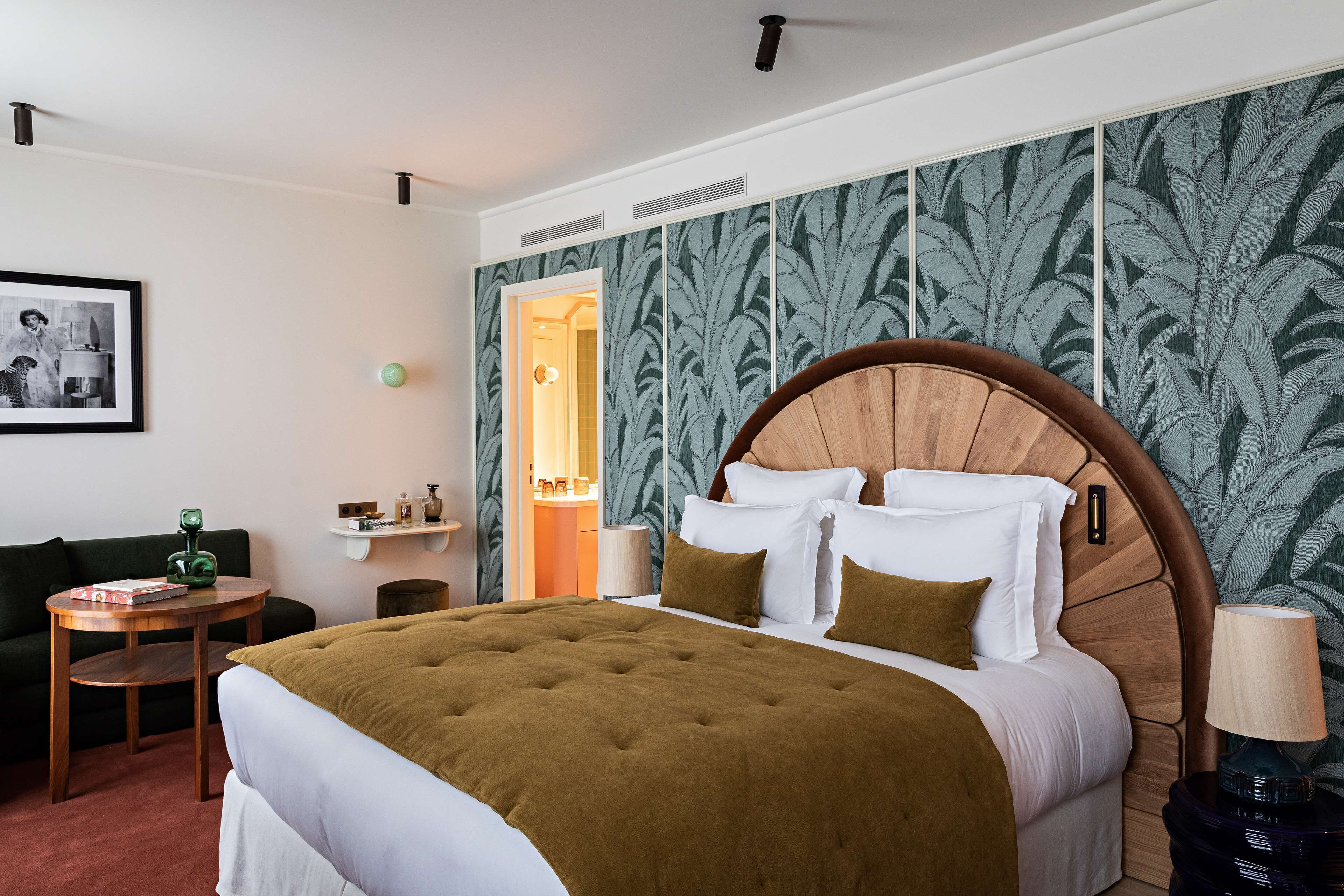 Quarto de hotel com uma cama grande, uma mesa pequena, uma cadeira verde, uma mesinha de cabeceira e uma parede com papel de parede estampado com folhas.