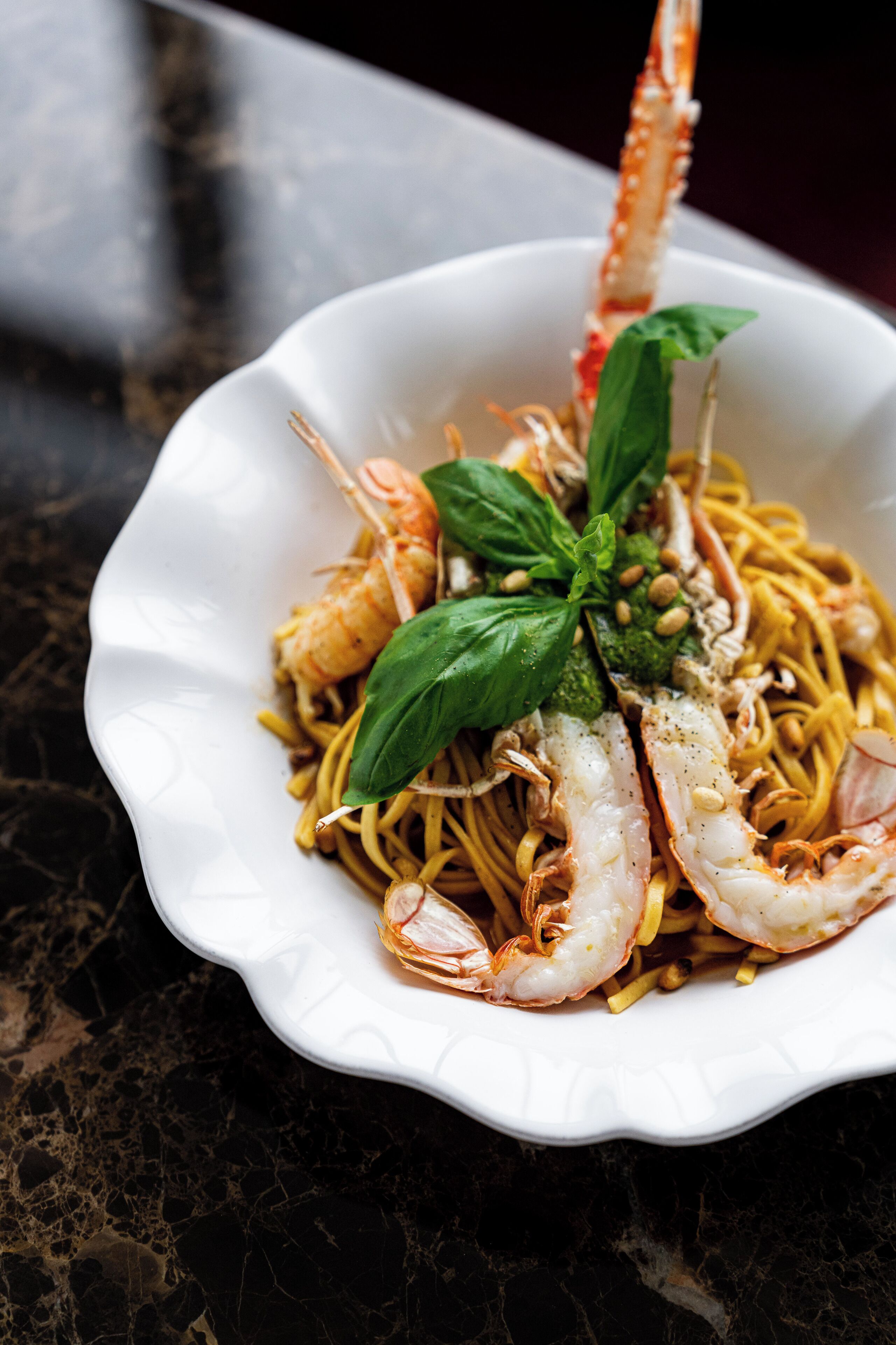 A white bowl of spaghetti with shrimp and basil on a dark marble surface.
