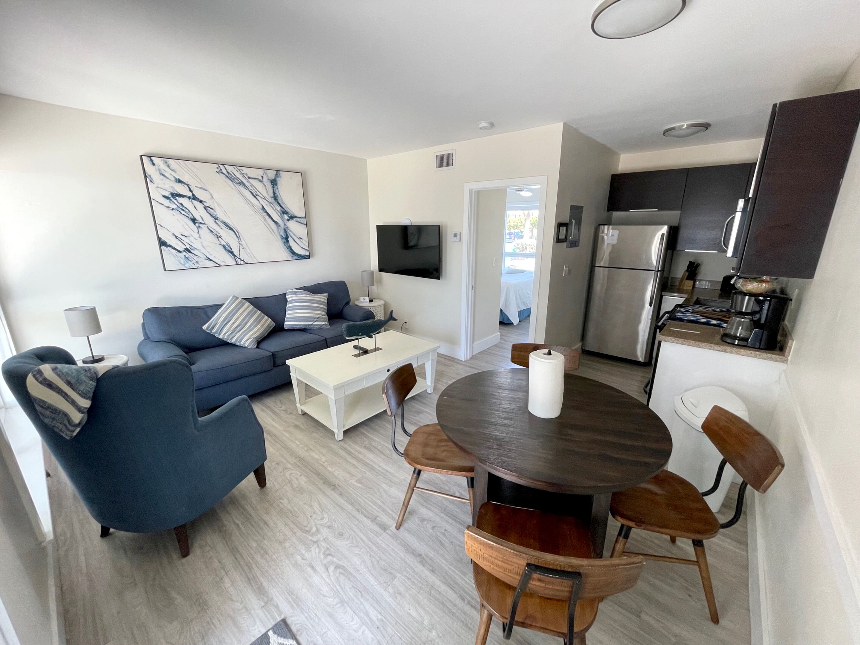 A modern living room with a blue sofa, a white coffee table, and a dining table with wooden chairs. A large painting is on the wall, and a kitchen with a refrigerator and stove is visible in the background.
