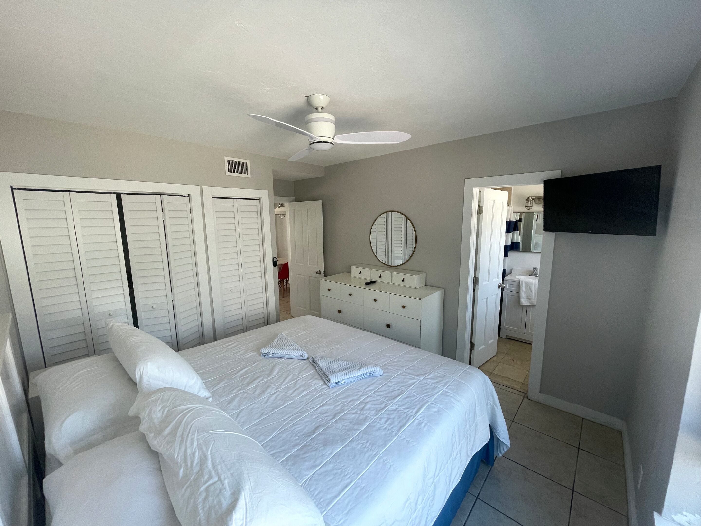 A bedroom with a bed, a dresser, a mirror, a ceiling fan, and a TV.