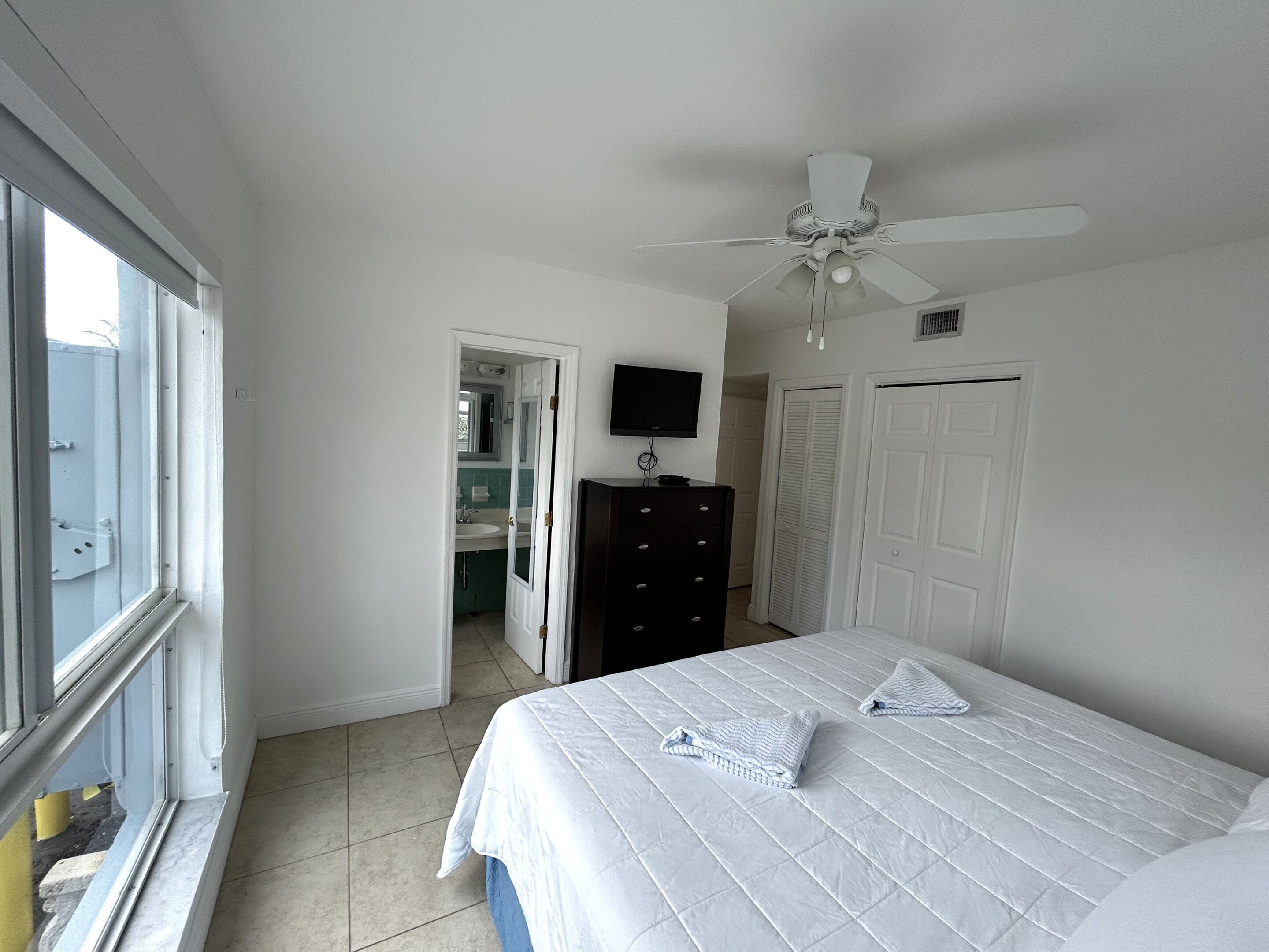 A bedroom with a bed, a ceiling fan, a dresser, a TV, and a bathroom visible through an open door.