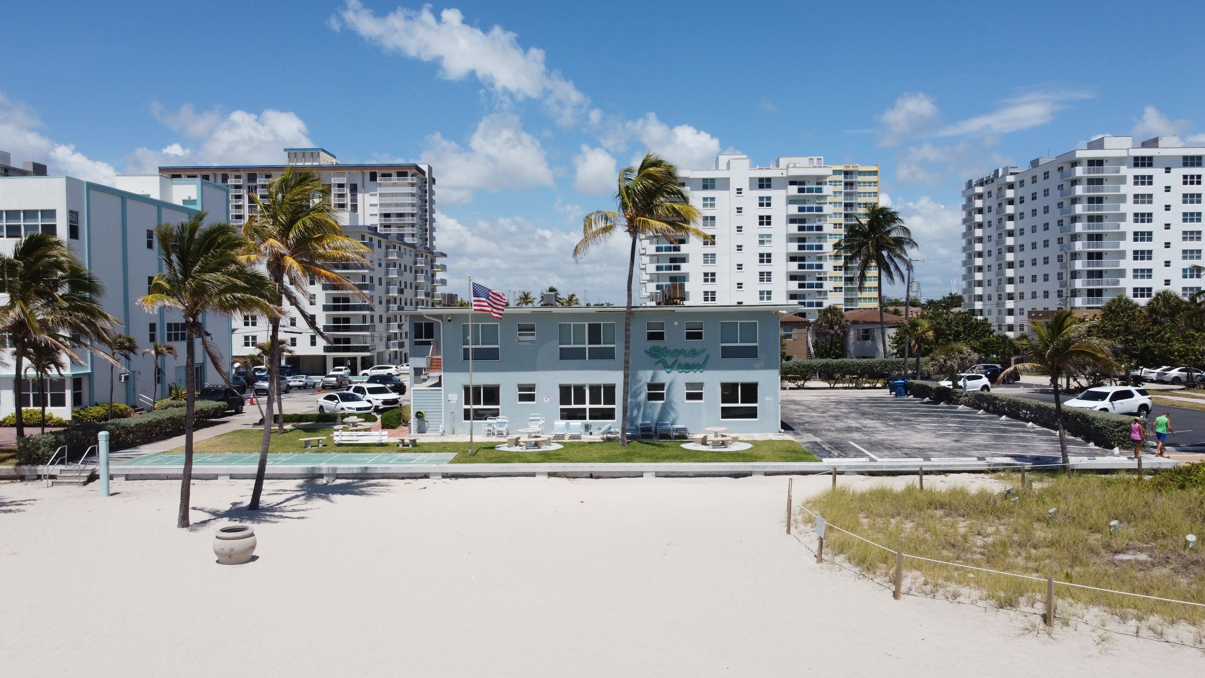 Ein Strandhotel mit mehreren Etagen, einem Sandstrand mit Palmen und einem Parkplatz.