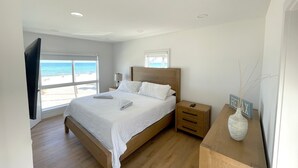 Une chambre dotée d’un grand lit, d’une tête de lit en bois, de tables de chevet, d’une commode, d’un téléviseur à écran plat fixé au mur et offrant une vue sur la plage par le biais d’une grande fenêtre.