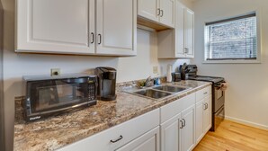 Una cocina con armarios blancos, encimera de granito, microondas, cafetera, pileta y ventana con persianas.