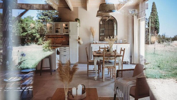 Un intérieur chaleureux et rustique, avec un coin repas en bois, une cuisine dotée de meubles blancs et une grande fenêtre donnant sur un jardin.