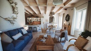 Un salon chaleureux avec un canapé bleu, une table basse en bois et des fauteuils en osier. On aperçoit une cuisine à l’arrière-plan, avec un coin repas.