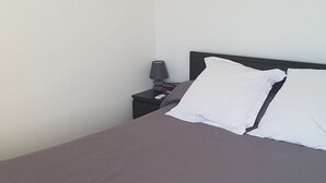 Una cama bien tendida con una manta gris y almohadas blancas, una mesita de noche con una lámpara y un cuadro en la pared.
