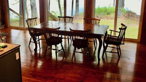 Uma ampla área de refeições com mesa e cadeiras de madeira, grandes janelas com vista para árvores e paisagem natural.