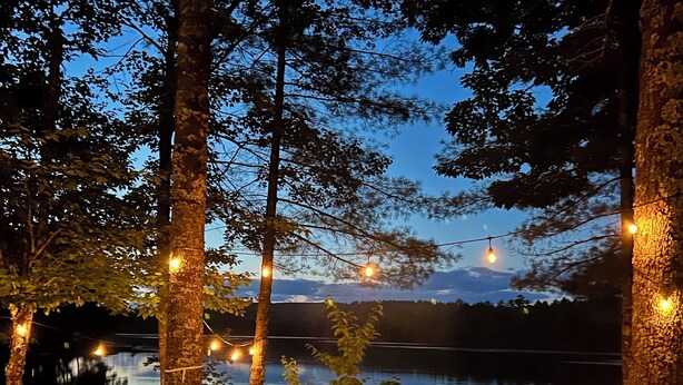 Fogueira rodeada por árvores, luzes de corda, um lago calmo e um céu limpo.