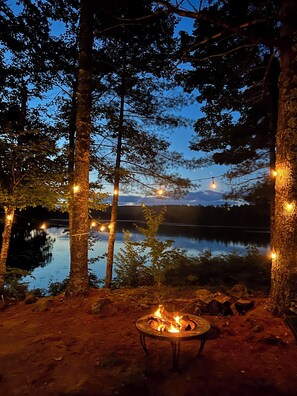 A campfire surrounded by trees with string lights, a calm lake, and a clear sky. - STUNNING WATERFRONT HOME ON PITCHER POND - LINCOLNVILLE. 3 bedrooms, 3 baths! (Lincolnville)