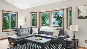 Un salon moderne avec un canapé d’angle, une table basse et un tapis à rayures. De grandes fenêtres offrent une vue sur les arbres et un immeuble.