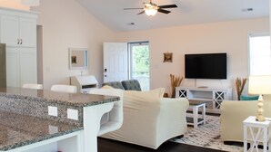 A modern living room with a kitchen area, a ceiling fan, a flat-screen TV, and a dining table. - Hale Aloha: An Aloha-Inspired Escape in the OBX (Kill Devil Hills)