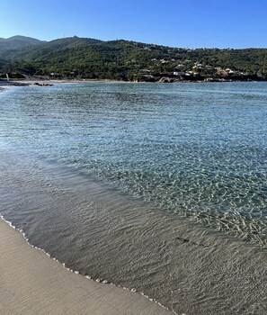 Ein Strand mit klarem Wasser, sandigem Ufer und hügeliger Landschaft im Hintergrund.