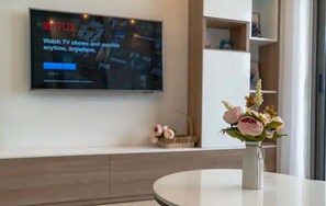 A modern living room with a flat-screen TV mounted on the wall displaying a Netflix advertisement. There is a round white table with a vase of flowers in the foreground.